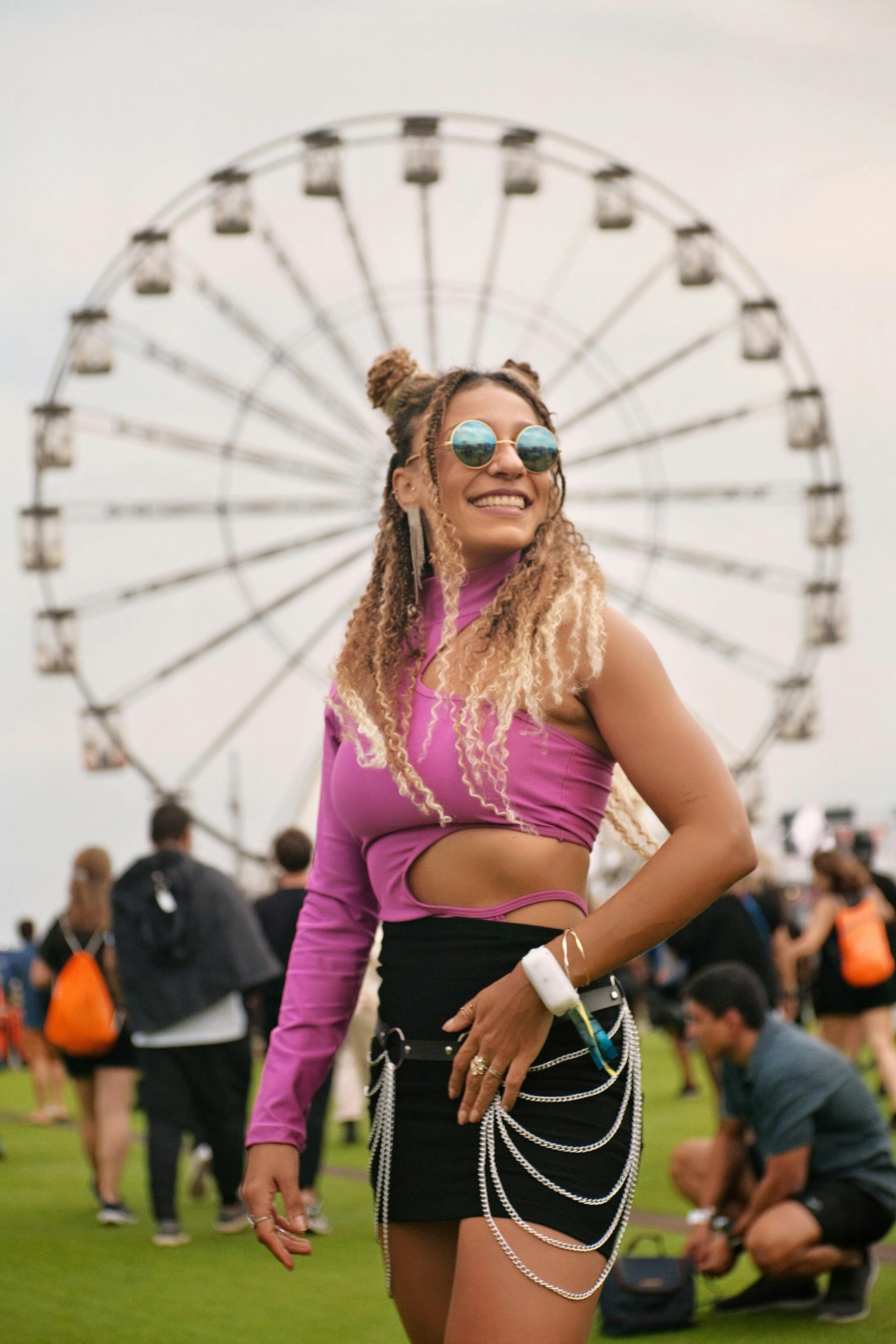 A smiling festivalgoer in a pink cut-out top and black mini skirt with layered silver chains poses in front of a Ferris wheel