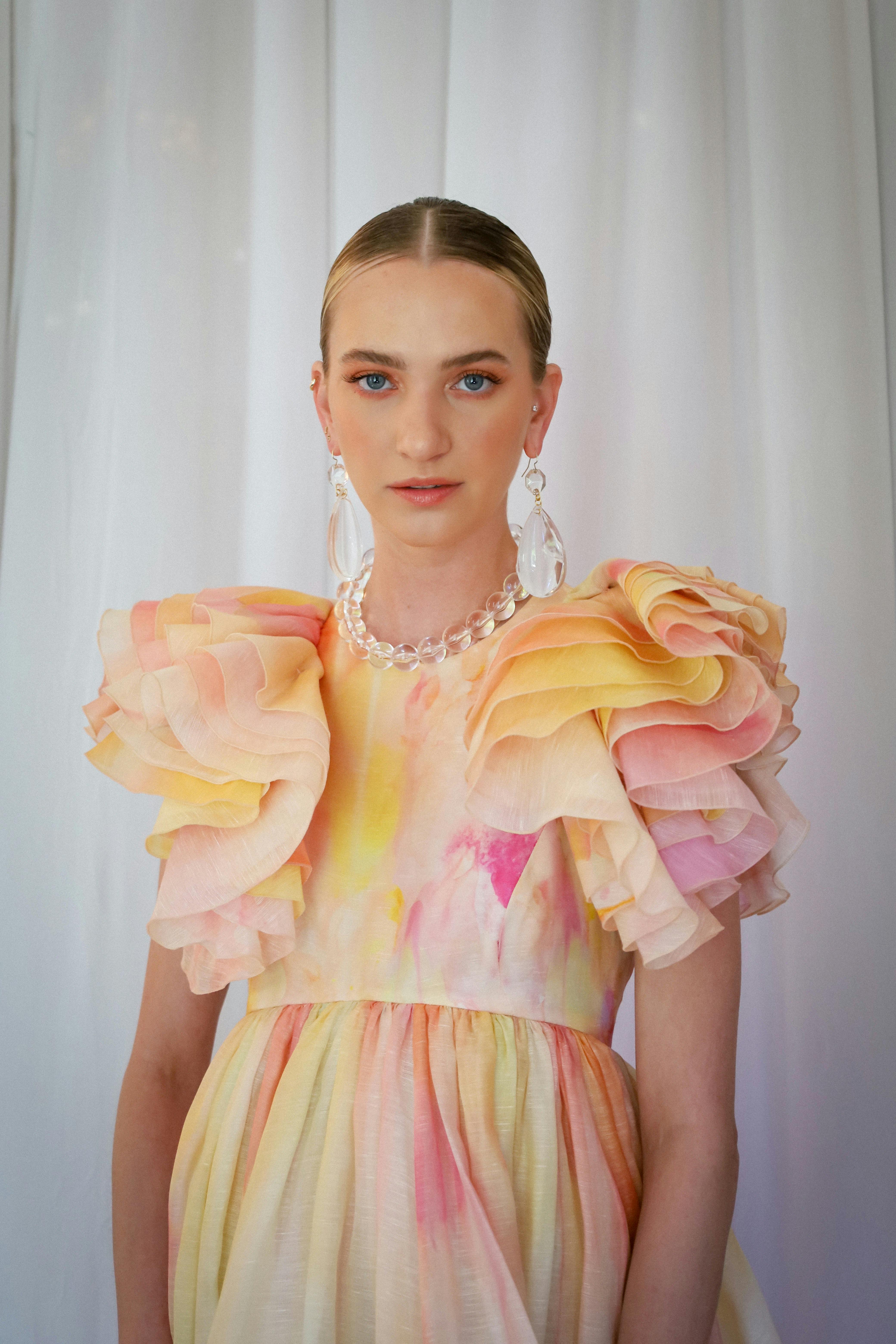 Model in a pastel watercolor-print dress with dramatic ruffled shoulders, paired with clear teardrop earrings and a beaded necklace.
