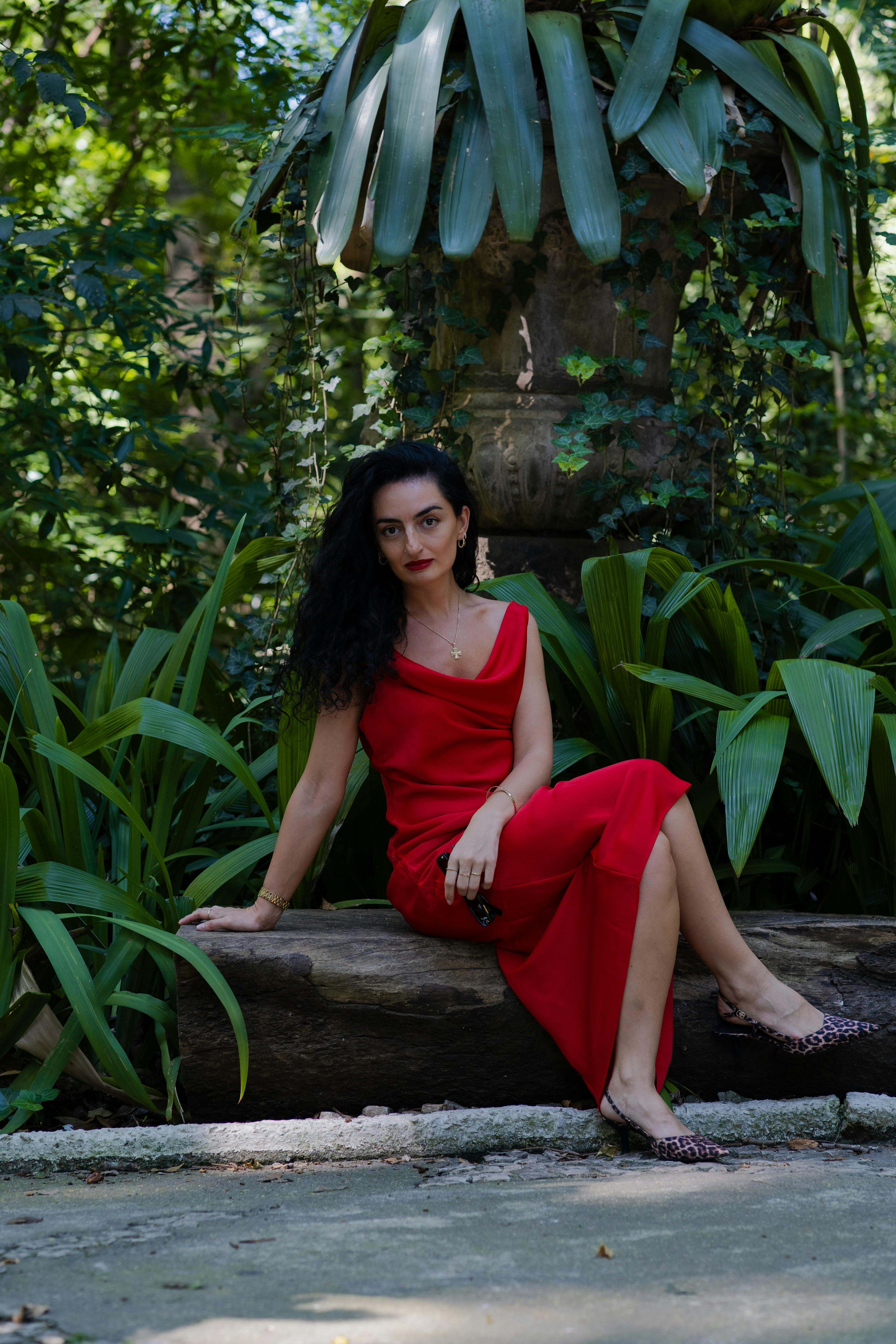 Woman in a red cowl-neck dress and leopard print heels seated on a log in a lush green garden.