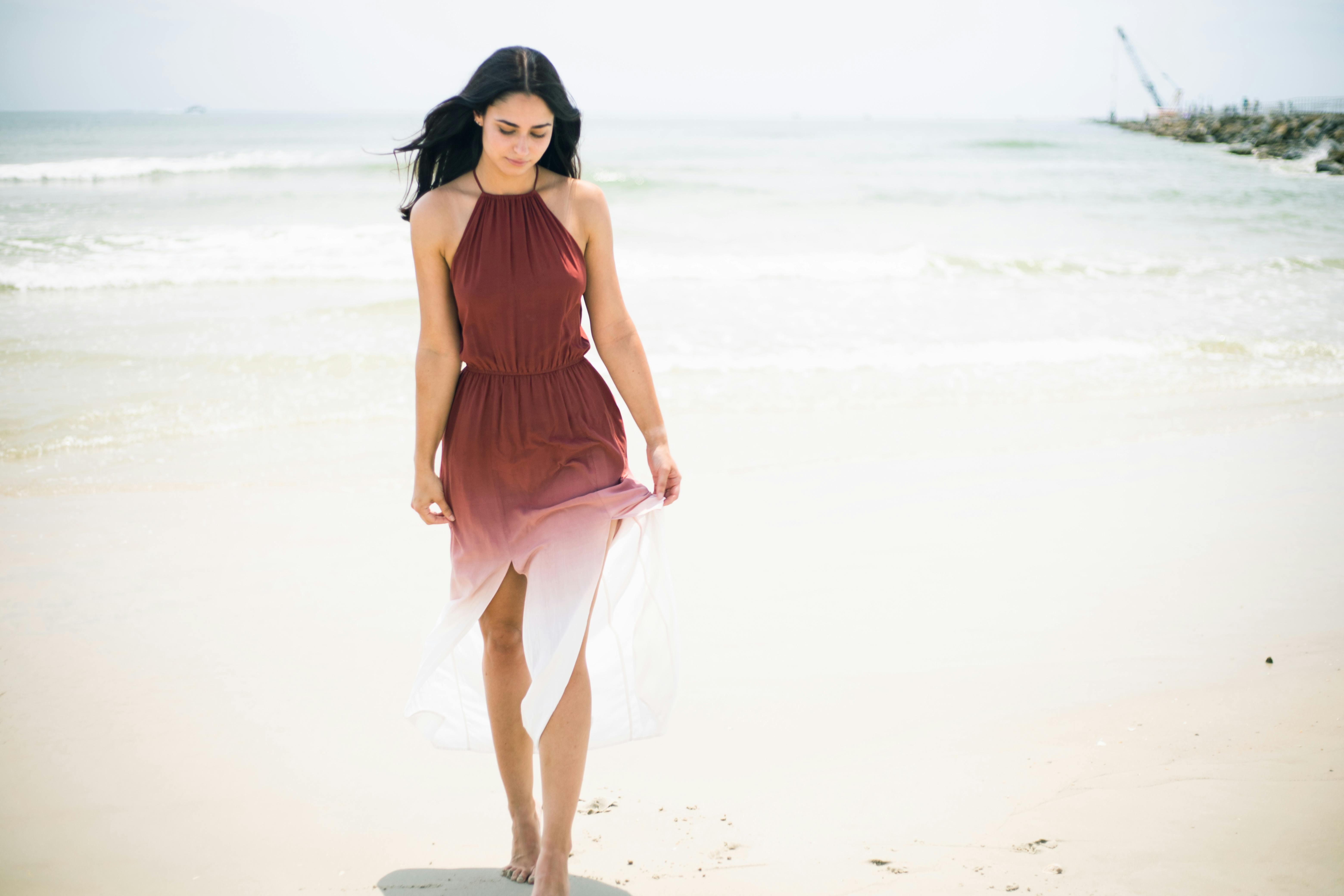 Woman in a burgundy ombré halter dress walking barefoot along the shoreline of a sandy beach.