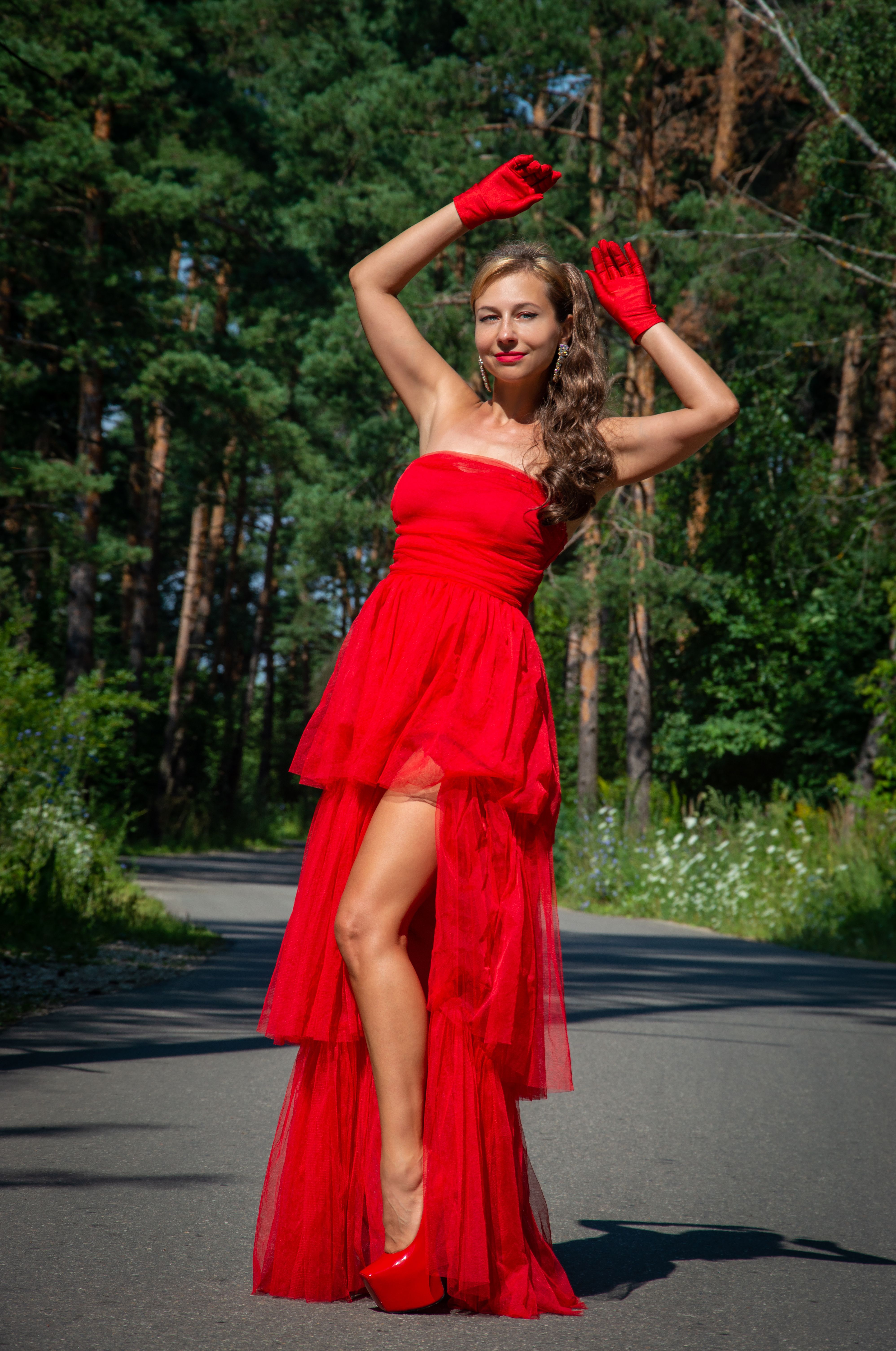 A woman in a tiered red tulle high-low gown with red opera gloves and red heels poses on a tree-lined road.