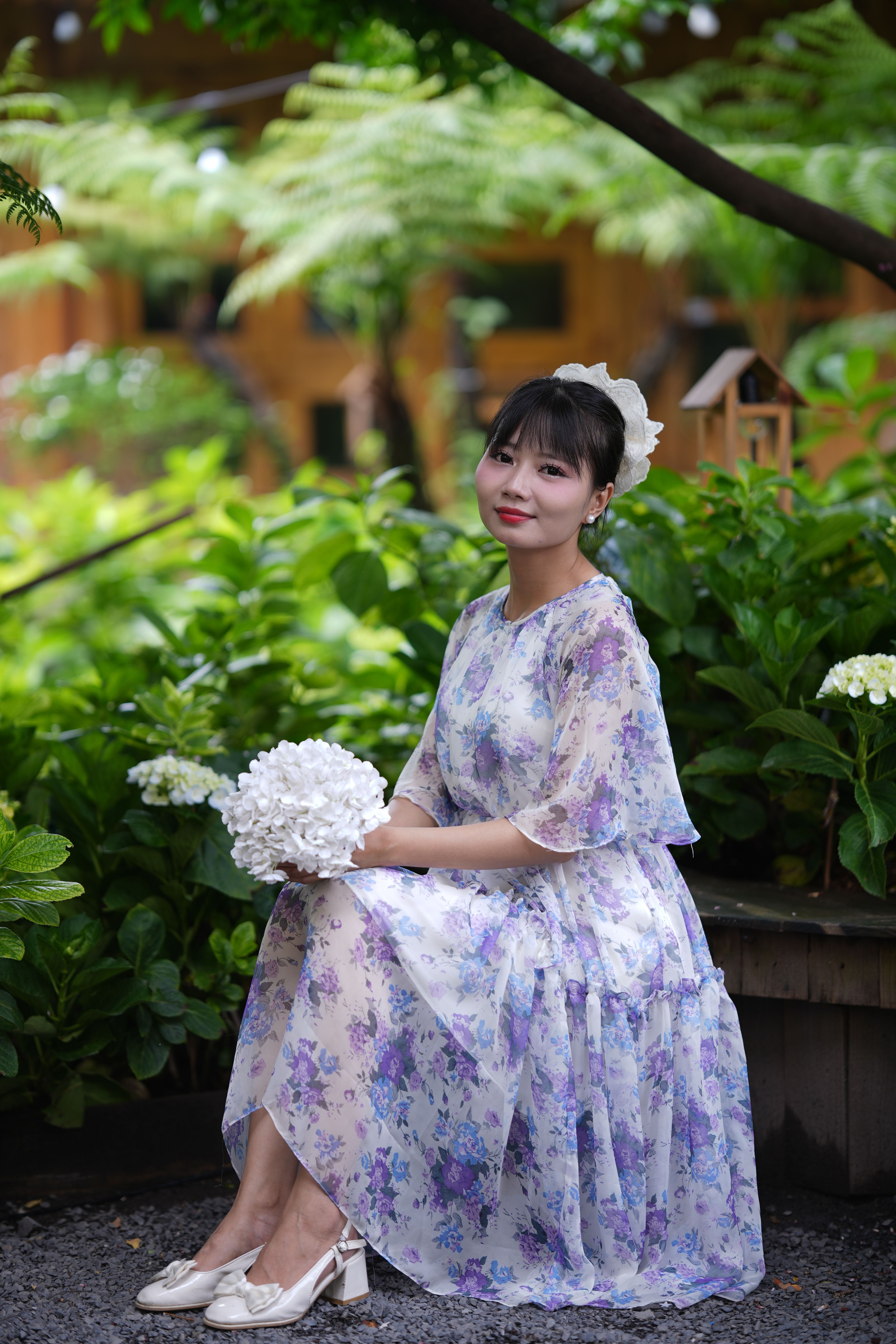 A woman in a lilac floral chiffon midi dress and white lace fascinator holds white hydrangeas in a garden