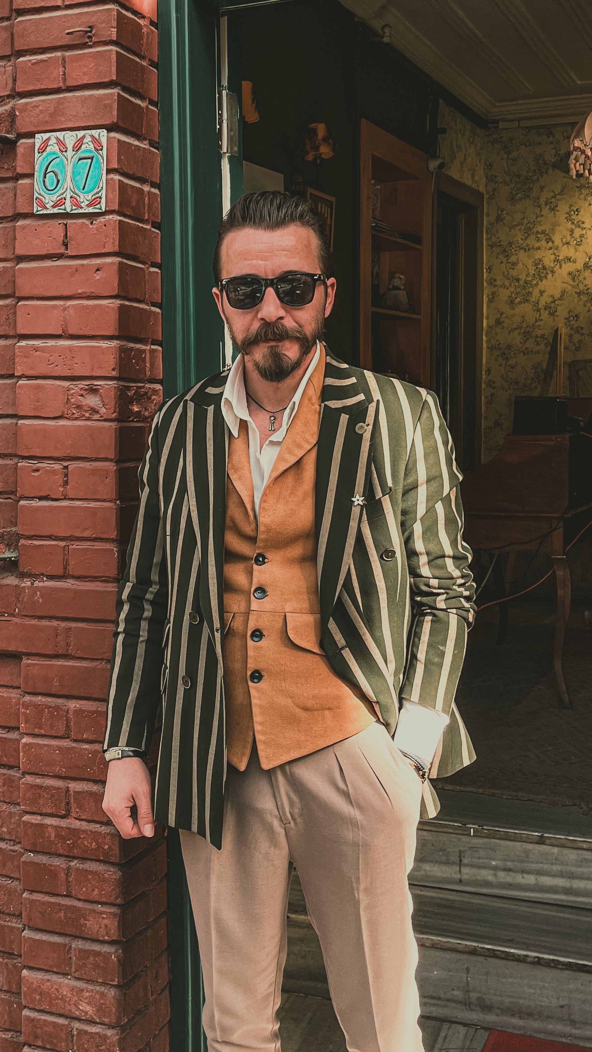 A man in a green-and-cream striped blazer over a tan suede waistcoat, open-collar shirt and cream trousers